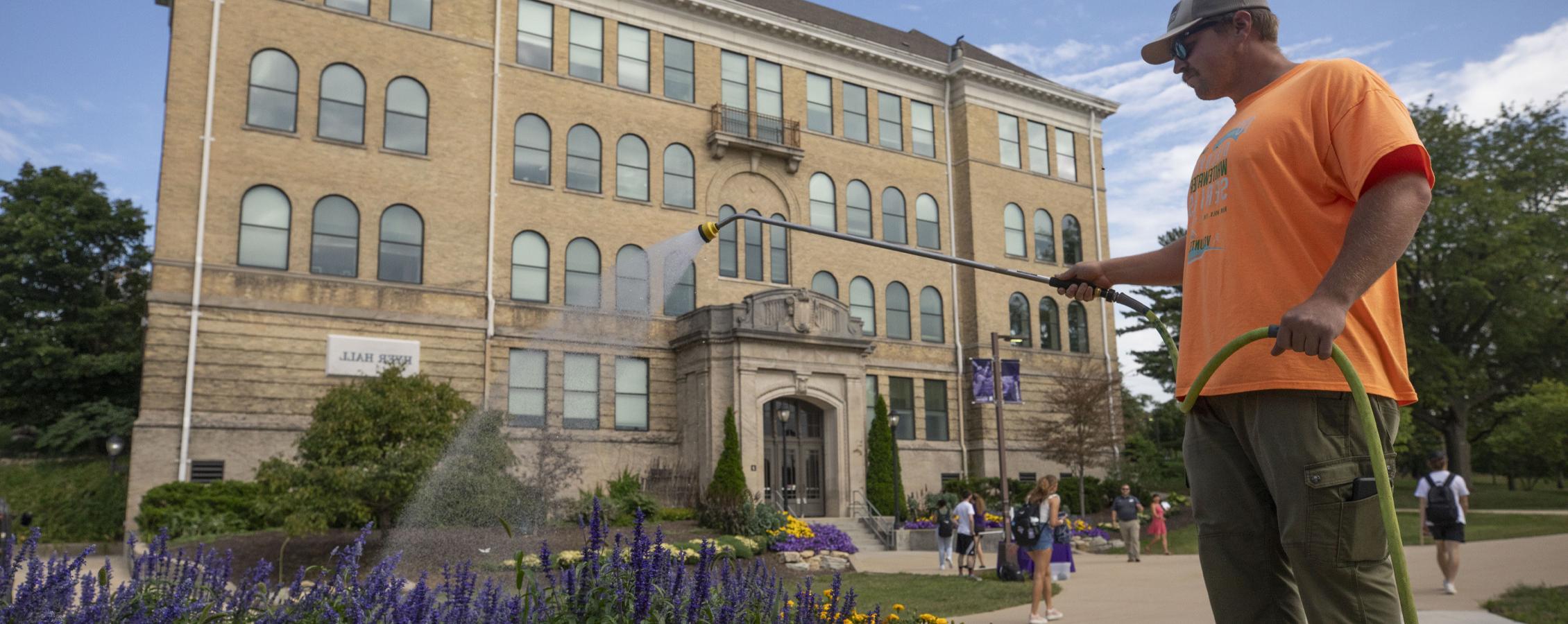 A person waters purple flowers in front of Hyer Hall.